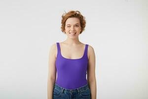Portrait of happy funny young attractive short-haired girl, winks and bites lip, wears in purple shirt, stands over white background. photo