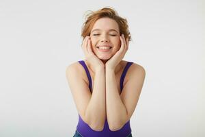 Close up of young nice short-haired lady wears in purple shirt and jeans, closes eyes and dreams about new dress, touches his cheeks and over white background. photo