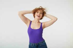 Portrait of young happy cute short-haired girl wears in purple shirt dancing with arms raised, touches hair, enjoying music and life, sings song, stands over white background. photo