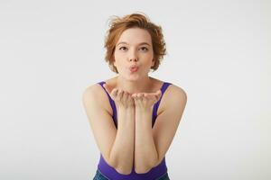 Close up of young cute short-haired girl wears in purple shirt and jeans, shows air kiss, protrudes palms at camera, expresses love to someone on distance, stands over white background. photo