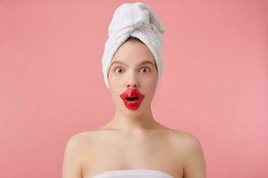Close up of young dazed lady after spa with a towel on her head, looks at the camera, with wide open eyes and mouth, patch for lips, stands over pink background. photo