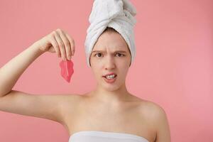 cerca arriba de ceñudo joven mujer con un toalla en su cabeza después ducha, participación un parche para labios, mira desconectado, soportes terminado rosado antecedentes. foto