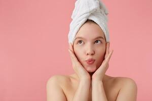 Close up of young happy woman after shower with a towel on her head, dreamily looking away with palms on cheeks, standing over pink background. photo