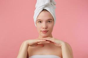 retrato de joven bonito mujer con natural belleza después ducha con un toalla en su cabeza, sonriente, mirando a el cámara terminado rosado antecedentes. foto