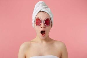 Wow Cool news Young happy amazed girl after spa with a towel on her head and mask on eyes, with wide open mouth and eyes, hears cool news, stands over pink background. photo