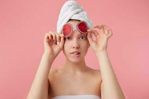 Close up of young confused lady with mask for eyes, after shower with a towel on her head, looks at the camera, bites lop and stands over pink background. photo