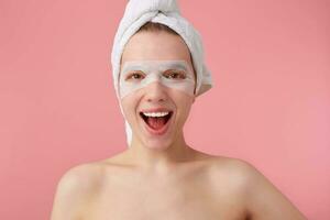 Portrait of young broadly smiling happy woman after spa with a towel on her head, with mask for eyes, enjoys for time for self care, stands over pink background. photo