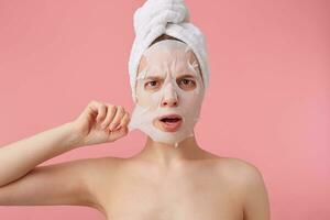 Close up of young disconnected woman with a towel on her head after shower, trying to remove the fabric mask from the face, stands over pink background. photo