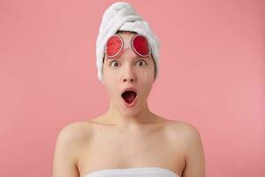 Wow Unbelievable news Young amazed girl after spa with a towel on her head and mask for eyes, with wide open mouth and eyes, hears new gossip, stands over pink background. photo