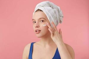 retrato de joven bonito mujer con natural belleza después ducha con un toalla en su cabeza, sonriente, mirando a el cámara terminado rosado antecedentes y pone en cara crema. foto