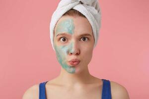 Portrait of young happy woman after shower with a towel on her head, with half face mask, surprised looking at the camera over pink background. photo