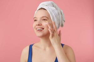 Close up of young nice woman with natural beauty with a towel on her head after shower, smiling, looking away over pink background and puts on face cream. photo