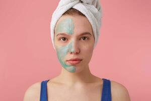 Close up of young nice lady with a towel on her head after shower, put a mask on the floor to compare the effect, looks at the camera stands over pink background photo