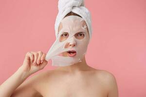 Close up of young woman with a towel on her head after shower, trying to remove the fabric mask from the face, stands over pink background. photo