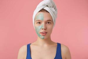 Photo of young nice lady with a towel on her head after shower, put a mask on the floor to compare the effect, looks at the camera stands over pink background