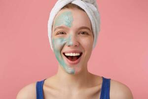 Portrait of young nice girl with half face mask, with a towel on her head after shower, smiling and looking at the camera, standing over pink background. photo