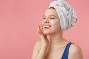 Photo of young nice joyful lady with natural beauty with a towel on her head after shower, stands over pink background and puts on face cream, looks away.