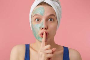 Close up of young woman after shower with a towel on her head, with half face mask, looking at the camera,showing a sign of silence gesture putting finger in mouth, stands over pink background. photo