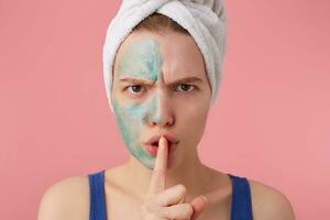 young frowning woman after shower with a towel on her head, with half face mask, looking at the camera,showing a sign of silence gesture putting finger in mouth, stands over pink background. photo