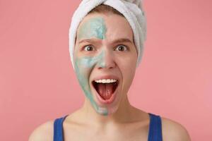 Close up of young shocked woman with a towel on her head after shower, with half face mask, looking at the camera and screaming, stands over pink background. photo