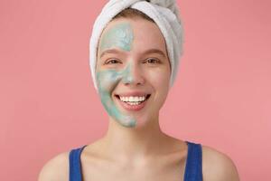 Portrait of young nice winked girl with half face mask, with a towel on her head after shower, smiling and looking away, touches cheek, standing over pink background. photo