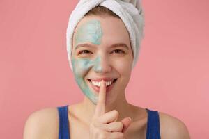 young smiling woman after shower with a towel on her head, with half face mask, looking at the camera,showing a sign of silence gesture putting finger in mouth, stands over pink background. photo