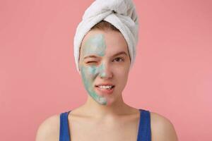 Portrait of young nice winked girl with half face mask, with a towel on her head after shower,smiling and looking at the camera over pink background. photo