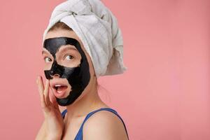 Portrait of young thinking woman after shower with a towel on her head, with black mask, touches face, stands over pink background. photo