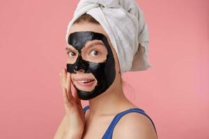 Close up of young thinking lady after shower with a towel on her head, with black mask, touches face, stands over pink background. photo