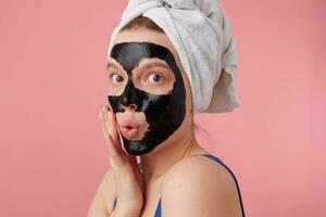 Portrait of young surprised woman after shower with a towel on her head, with black mask, touches face, with shocked expression on face, stands over pink background. photo