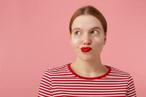 young mysterious red-haired lady with red lips and with patches under the eyes, wears in a red striped T-shirt, confused looks at the left side, something plotting, stands over pink background. photo