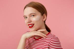 bonito sonriente Pelirrojo niña usa en un rojo a rayas camiseta, con rojo labios y con parches debajo el ojos, toques el cheen, soportes terminado rosado antecedentes y disfrutando gratis hora para piel cuidado. foto