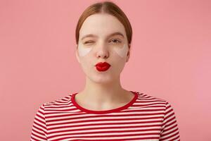 retrato de linda joven sonriente Pelirrojo mujer con rojo labios y con parches debajo el ojos, usa en un rojo a rayas camiseta, mira a el cámara y guiño, envía beso, soportes terminado rosado antecedentes. foto