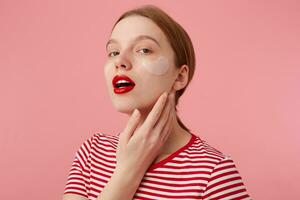Portrait of cute young red-haired girl with red lips and with patches under the eyes, wears in a red striped T-shirt, looks at the camera and touches the cheek, stands over pink background. photo