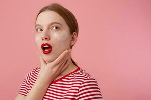 Portrait of cute thinking young red-haired girl with red lips and with patches under the eyes, wears in a red striped T-shirt, looks away, touches the cheek, stands over pink background. photo