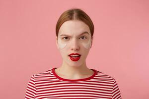 retrato de linda joven descontento Pelirrojo dama con rojo labios y con parches debajo el ojos, usa en un rojo a rayas camiseta, soportes terminado rosado antecedentes con amplio abierto boca y elevado ceja. foto