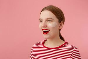 retrato de linda joven contento Pelirrojo dama con rojo labios y con parches debajo el ojos, usa en un rojo a rayas camiseta, mira lejos y en general sonriente, soportes terminado rosado antecedentes. foto