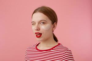 retrato de linda joven Pelirrojo mujer con rojo labios y con parches debajo el ojos, usa en un rojo a rayas camiseta, mira a el cámara y guiño, muestra lengua, soportes terminado rosado antecedentes. foto