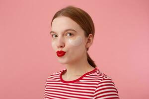 Portrait of cute young smiling red-haired girl with red lips and with patches under the eyes, wears in a red striped T-shirt, looks at the camera and sends kiss, stands over pink background. photo