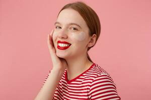 joven sonriente Pelirrojo dama con rojo labios usa en un rojo a rayas camiseta, con parches debajo el ojos, toques el mejilla, disfrutando gratis hora para piel cuidado y soportes terminado rosado antecedentes. foto