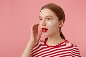 retrato de linda joven Pelirrojo niña con rojo labios y con parches debajo el ojos, usa en un rojo a rayas camiseta, mira lejos y piensa acerca de nuevo vestido, soportes terminado rosado antecedentes. foto