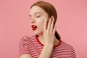 Young red-haired girl with red lips and with patches under the eyes, wears in a red striped T-shirt, touches the cheek, stands over pink background with closed eyes enjoying free time for skin care. photo
