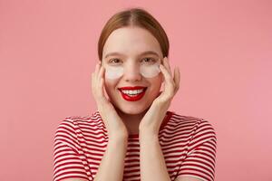 Cute young red-haired woman in a red striped T-shirt, with red lips , touches his face with fingers, very pleased with my new patches from dark circles under my eyes. Stands over pink background. photo