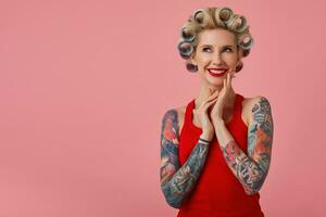 Cheerful young attractive blonde lady with tattooed hands smiling and looking dreamily upwards, foretasting upcoming party and preparing for it, standing against pink background photo