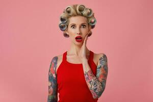 Open-eyed young pretty blonde woman with tattooes and festive makeup holding palm on her cheek and looking amazedly at camera, getting ready for upcoming party, isolated over pink background photo