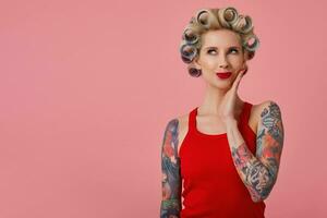 Indoor photo of beautiful positive young blonde lady with tattooes dressed in red shirt looking dreamily upwards with palm on her cheek, posing over pink background with curlers on her head
