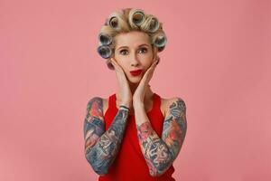 Beautiful young blonde tattooed lady with red lips holding her face with palms and looking positively at camera, having curlers on her head and preparing for going out, isolated over pink background photo