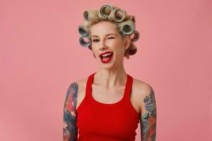Close-up of attractive young blonde tattooed woman making hairdo while preparing for holiday celebration, winking positively at camera while standing over pink background with curlers on her hair photo