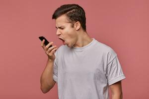 interior foto de enojado bonito joven oscuro peludo masculino en pie terminado rosado antecedentes con móvil teléfono en mano, teniendo desagradable hablar en teléfono y gritos con el ceño fruncido dentro auricular