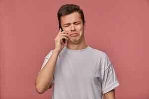 Studio photo of upset young dark haired man in casual clothes standing over pink background, holding smartphone in hand with sad face, hearing bad news on phone